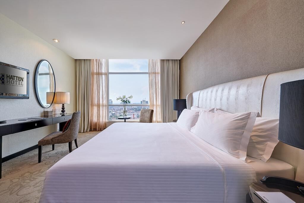 A hotel room with a large bed, a desk with a chair, a round mirror, and a view of the city through large windows.
