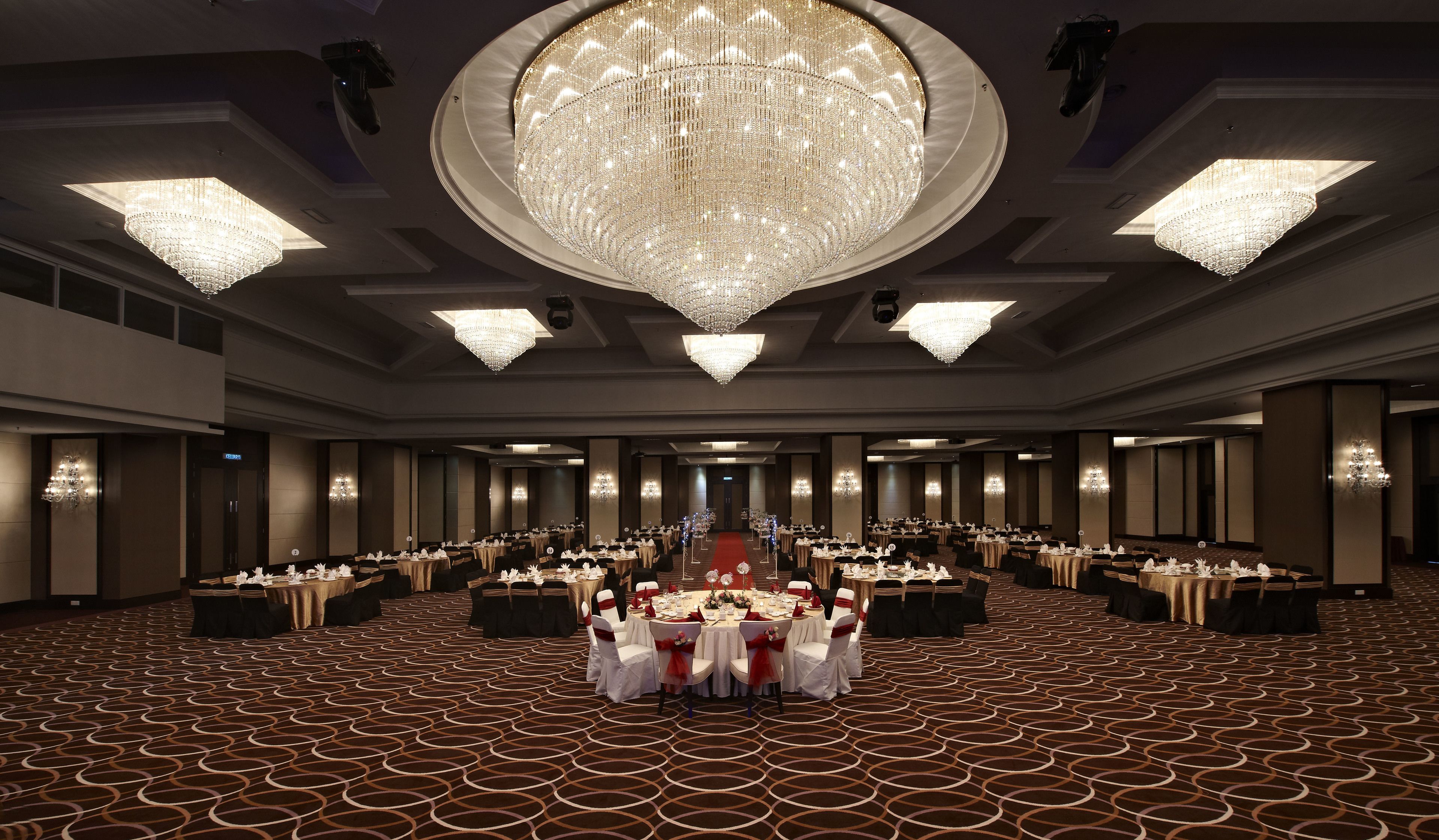 A large banquet hall with multiple round tables set for dining, a grand chandelier, and a patterned carpet.