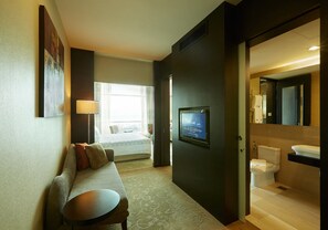 Habitación de hotel con sofá, una mesita, televisor, baño con inodoro y lavabo, y ventana con cortinas.