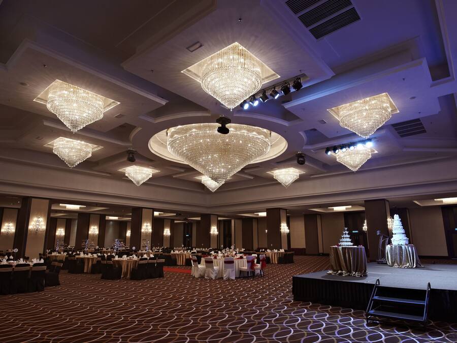 A banquet hall with multiple tables set for a formal event, large chandeliers, and a stage with a tiered cake.