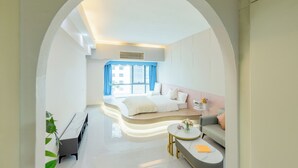 Moderni hotellihuone, jossa on kaareva sänky, sohva, pieni pyöreä pöytä ja ikkuna, jossa on verhot.