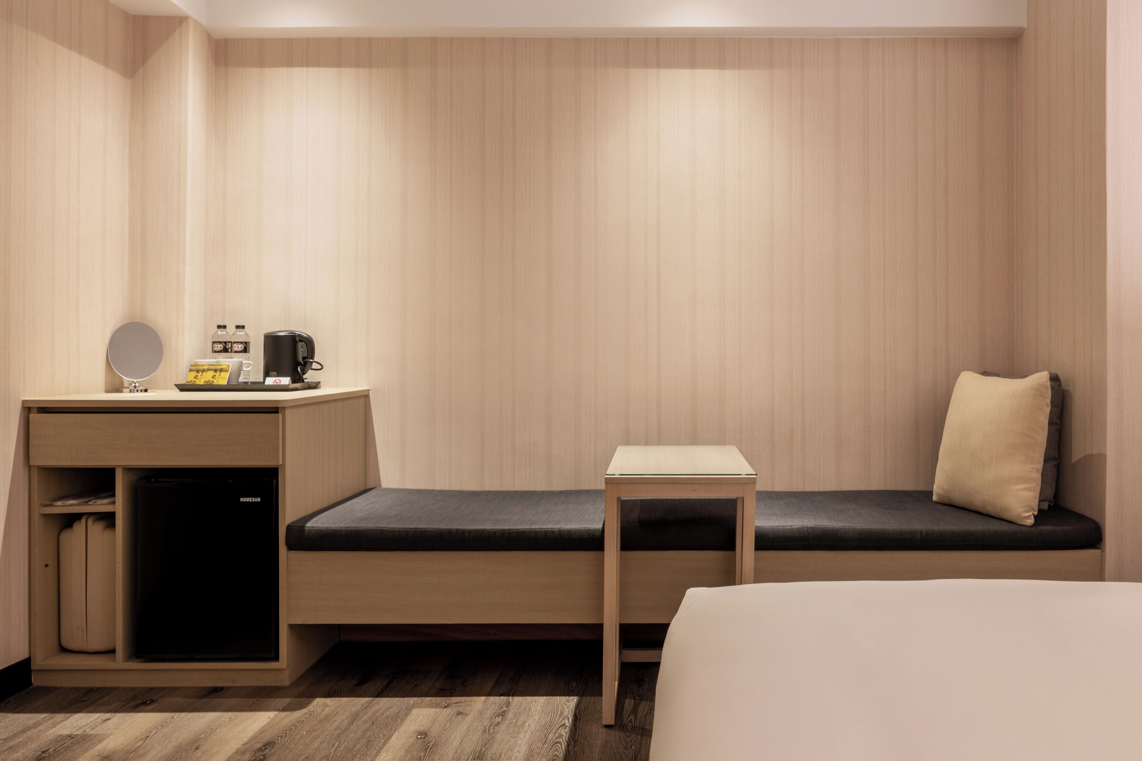 Habitación de hotel compacta con cama, un pequeño escritorio con cafetera y un banco con almohadón.