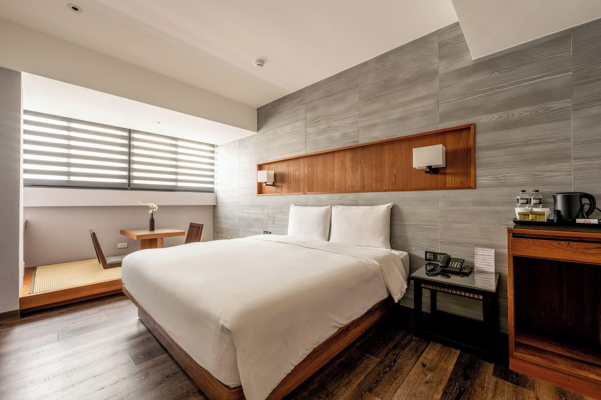 A modern hotel room with a large bed, wooden headboard, a small desk with a telephone, and a window with blinds.