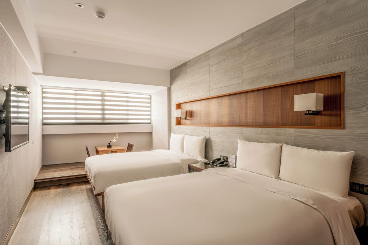 A modern hotel room with two beds, a wooden headboard, a small desk, and a window with blinds.