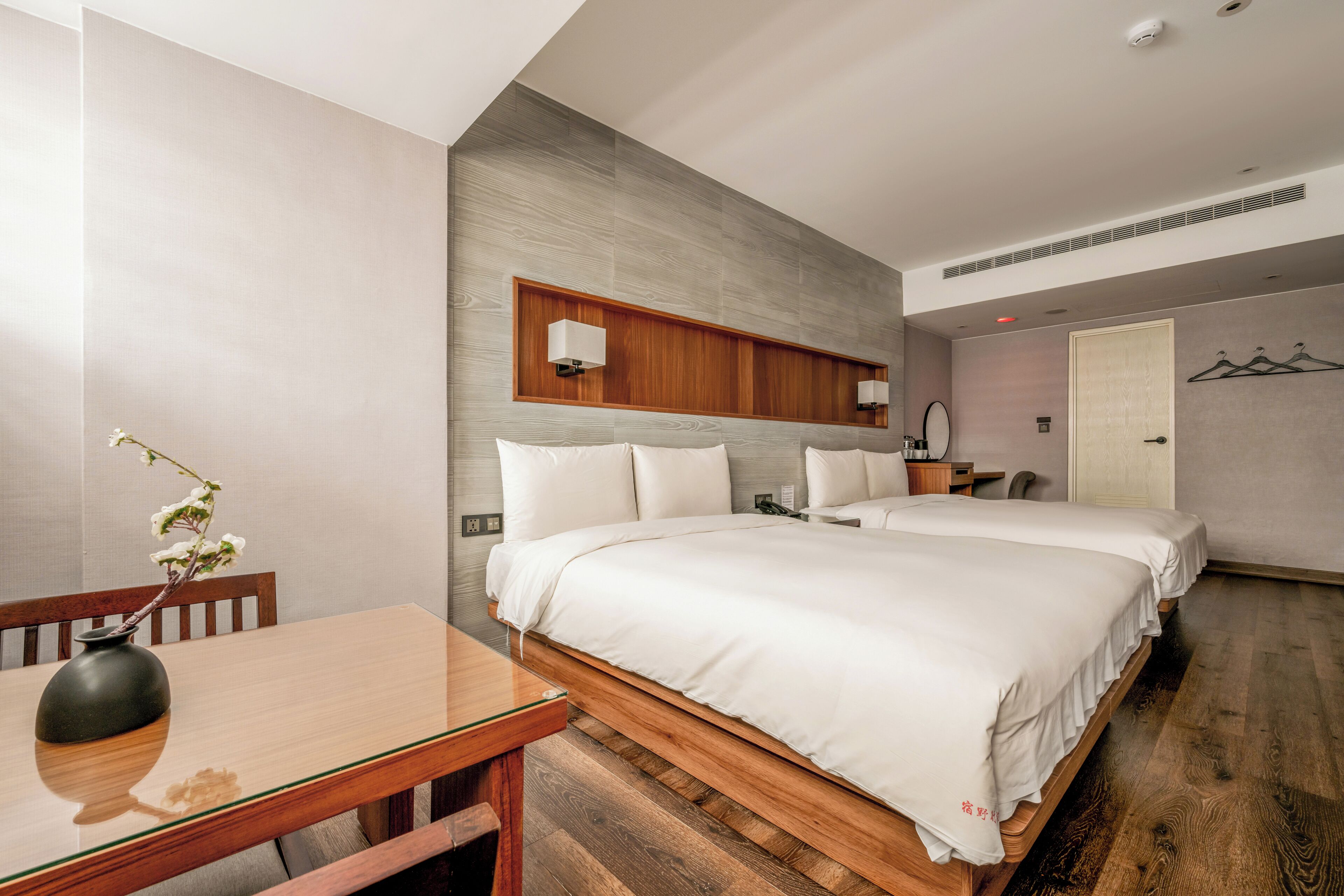 A modern hotel room with two beds, a wooden table, a vase with flowers, and a wall-mounted shelf with lamps.