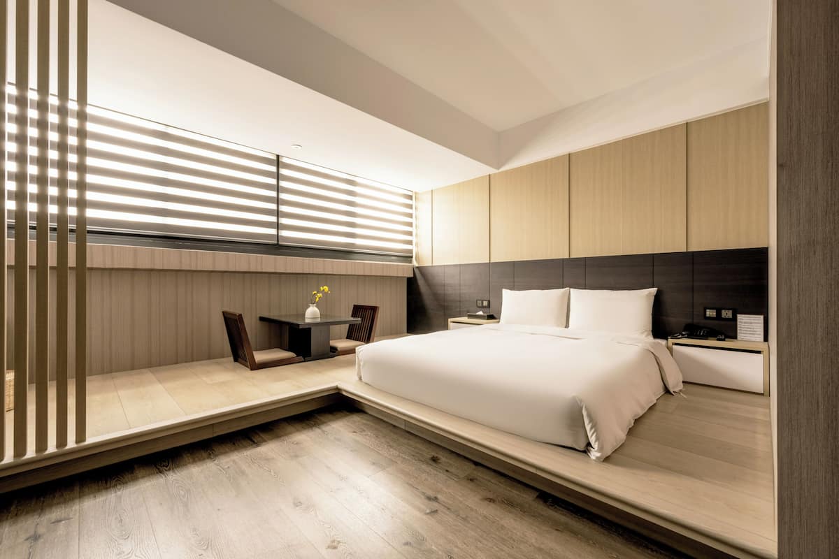 A modern bedroom with a large bed, bedside tables, a desk, and a window with blinds.