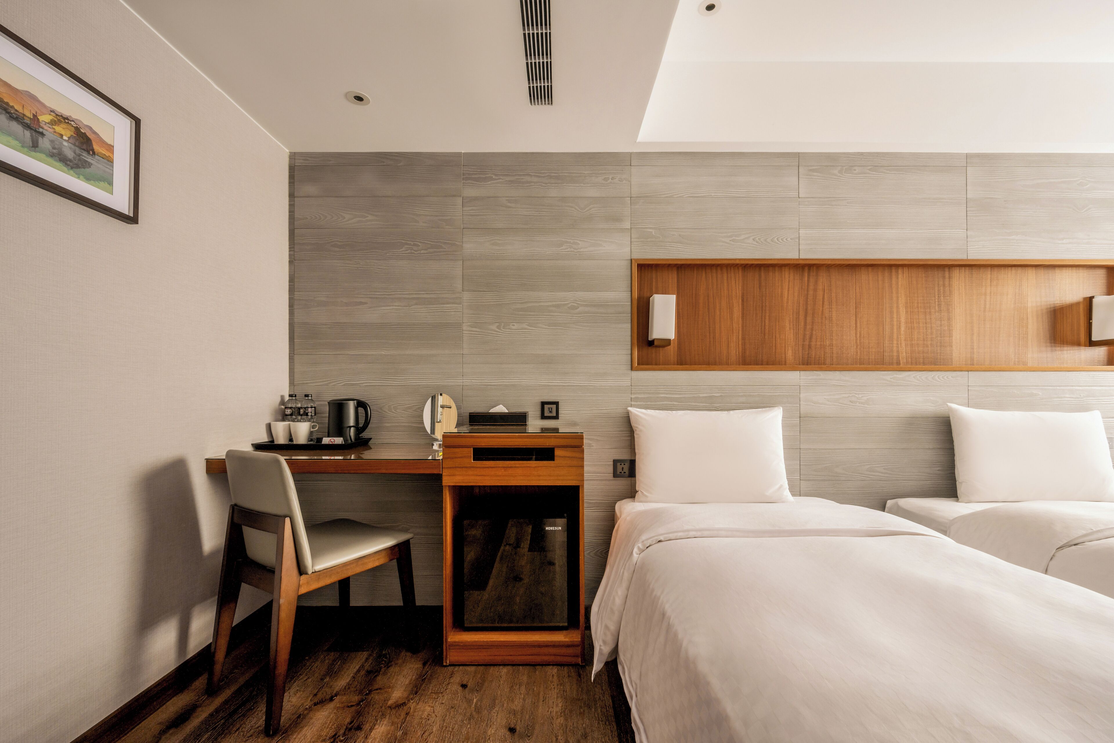 Een moderne hotelkamer met een houten bureau, een stoel, een bed met witte lakens en een kunstwerk aan de muur.