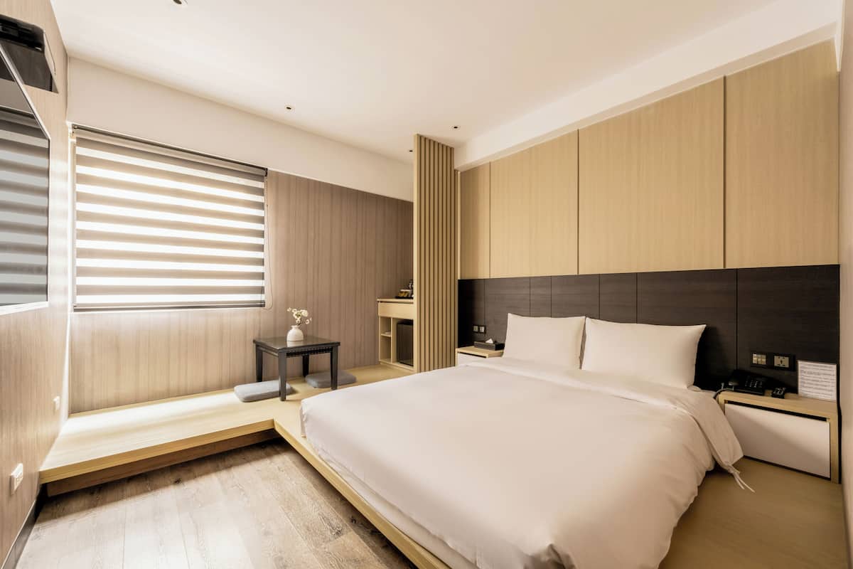 A modern hotel room with a large bed, a bedside table, a nightstand, and a window with blinds.