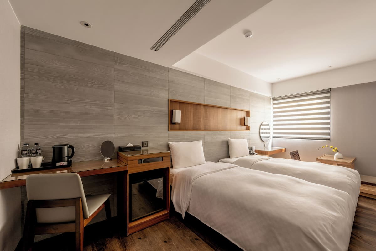 A modern hotel room with a wooden desk, two beds, a small seating area, and a large window with blinds.
