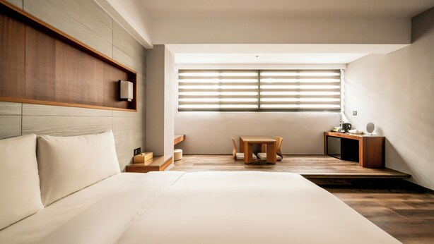 A modern hotel room with a large bed, a wooden headboard, a small table with chairs, a desk with a lamp, and a large window with blinds. - Suye Hotel (Taichung)