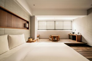 A modern hotel room with a large bed, a wooden headboard, a small table with chairs, a desk with a lamp, and a large window with blinds. - Suye Hotel (Taichung)