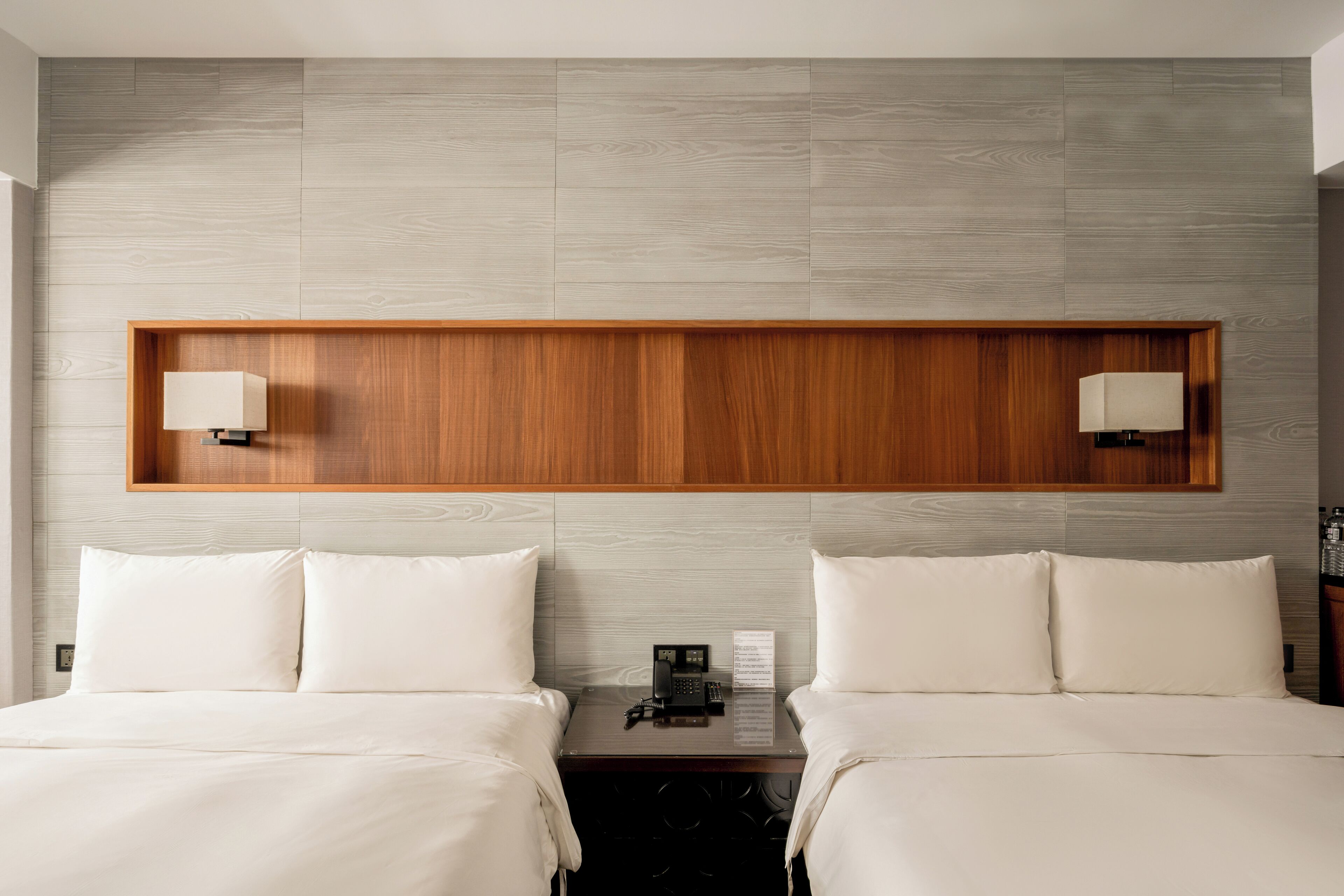 A hotel room with two beds, a wooden headboard, a nightstand with a lamp, and a telephone.