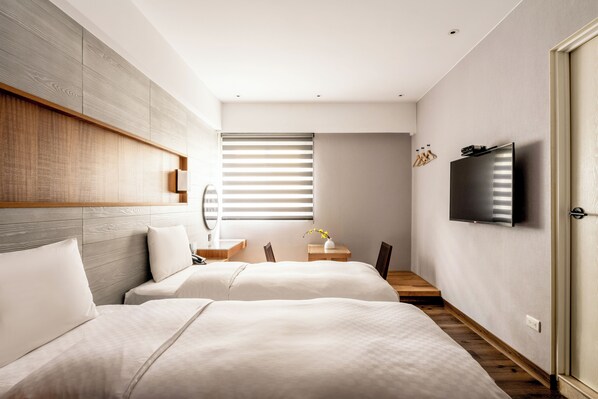A modern hotel room with two beds, a flat-screen TV, a wooden headboard, and a large window with blinds.