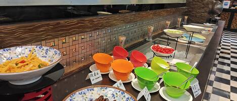 Buffet com diversos pratos em tigelas coloridas, um painel de azulejos e chão quadriculado.