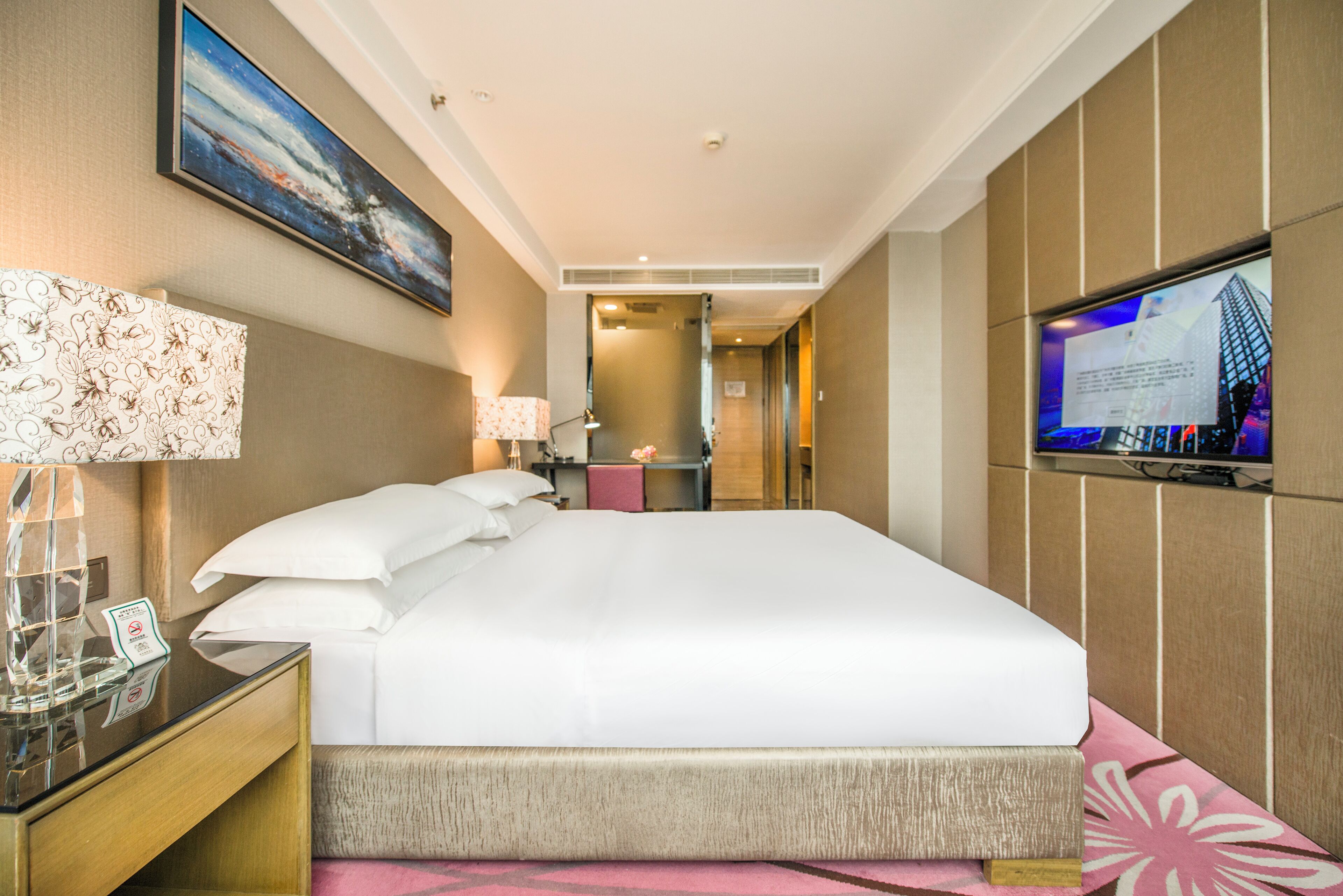 Habitación de hotel con una cama grande, un televisor montado en la pared, un escritorio con lámpara y una mesita con superficie de vidrio.