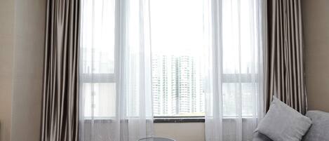 A hotel room with a bed, a small table, sheer curtains, and a view of tall buildings.