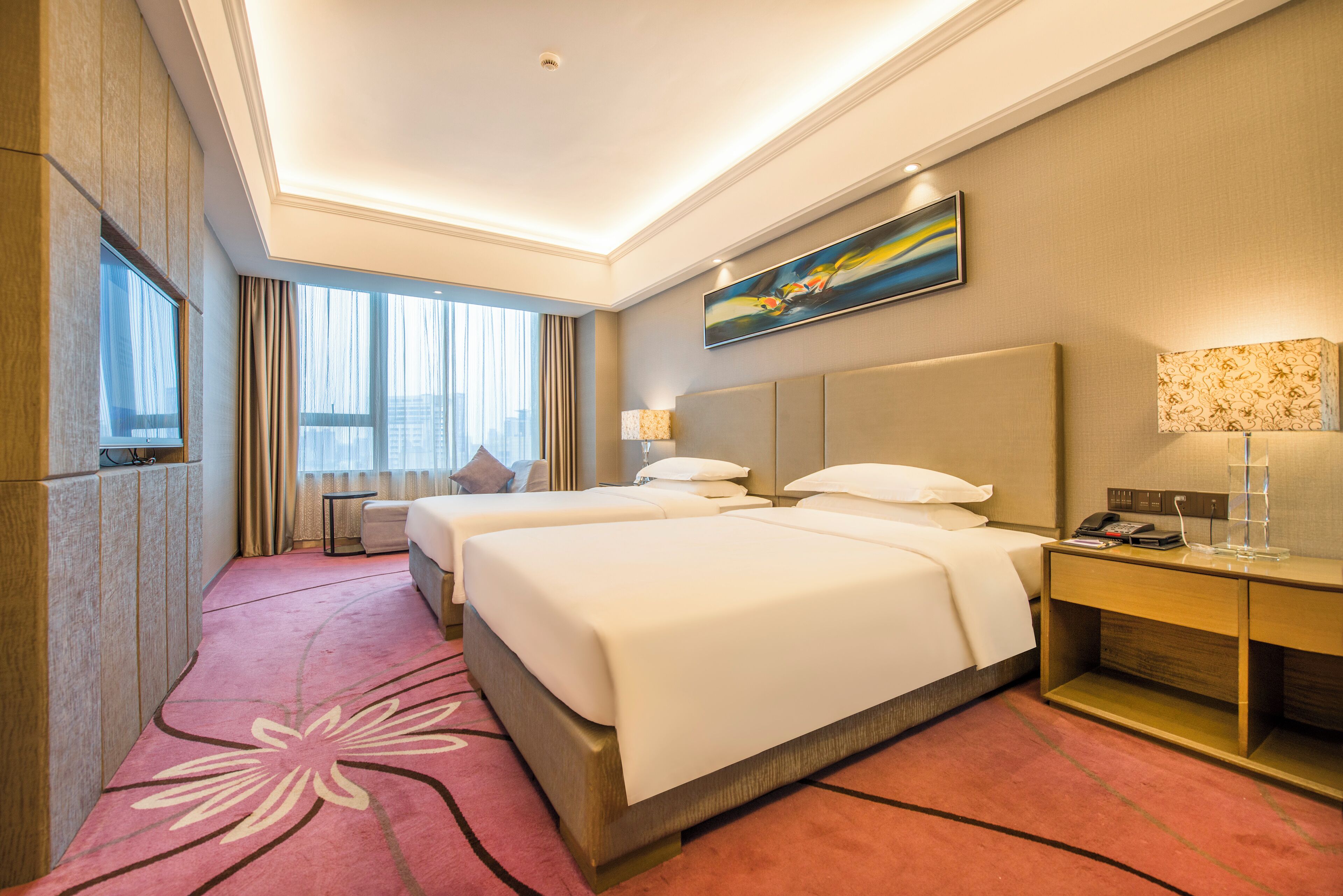 A hotel room with two beds, a desk, a lamp, a painting, and a large window with curtains.