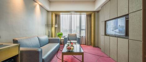 A modern hotel room with a sofa, armchair, coffee table, TV, and a view of the city through large windows.