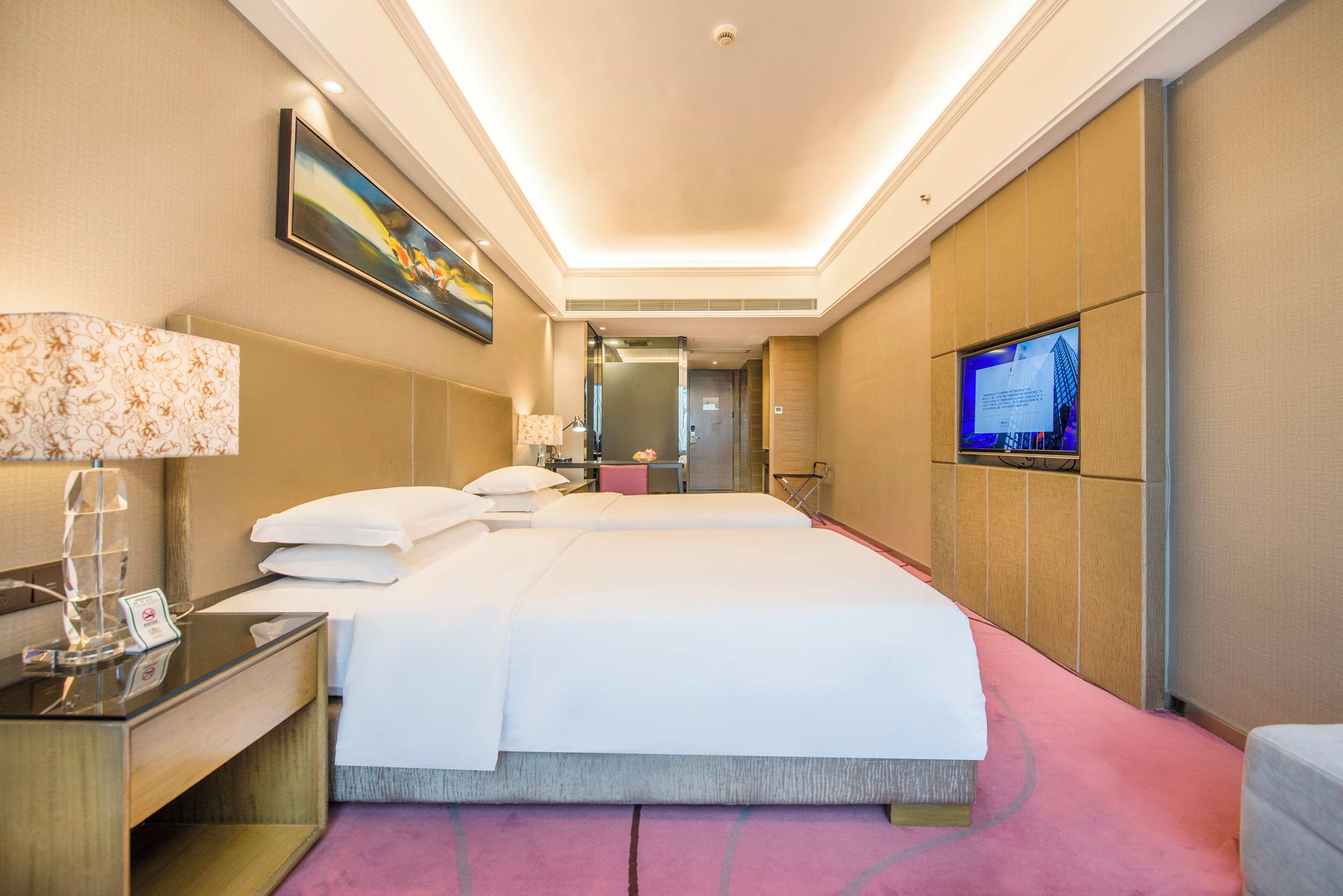 A hotel room with a large bed, a desk with a lamp, a television, and a painting on the wall.