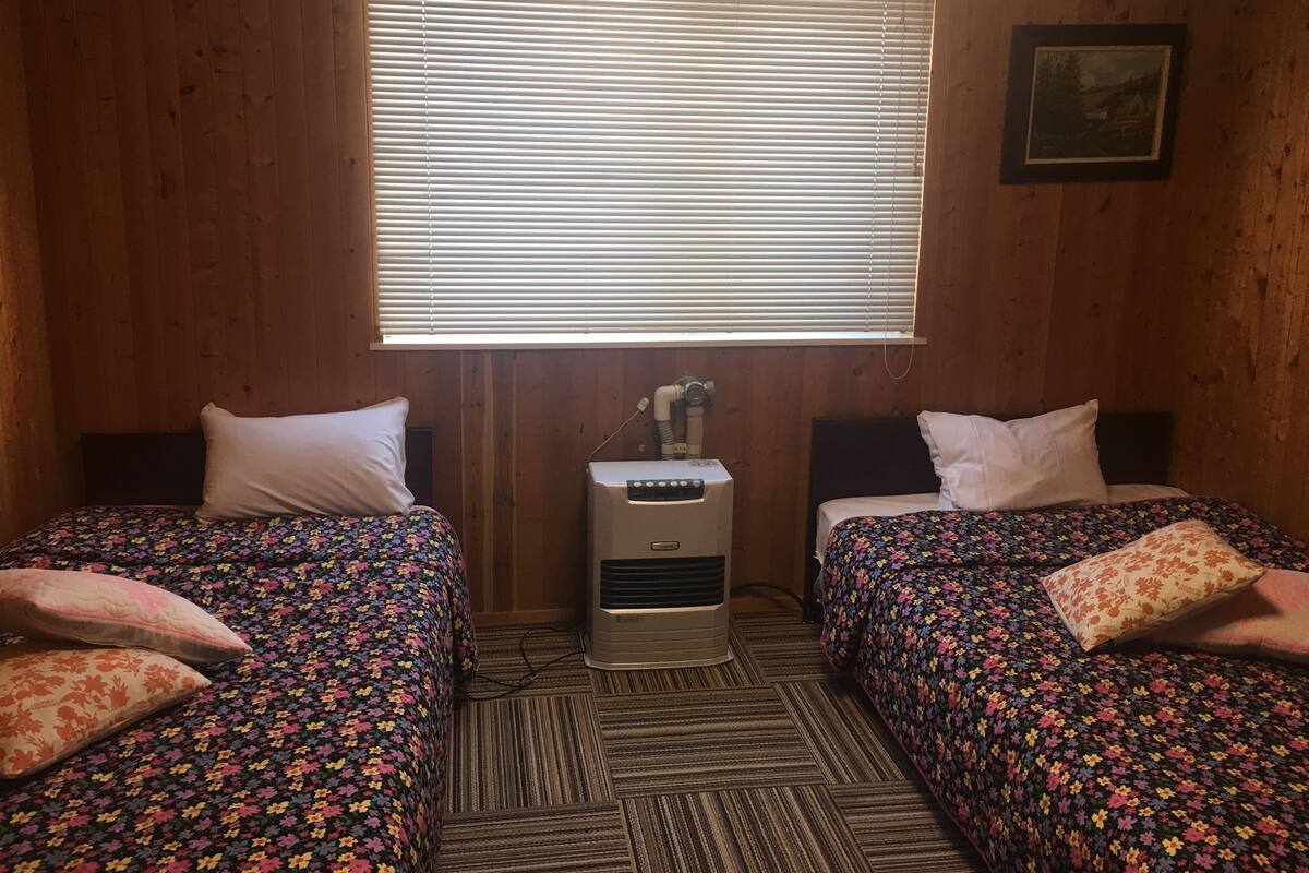 A room with two beds, each with floral bedding, a window with blinds, a heater, and a framed picture on the wall.