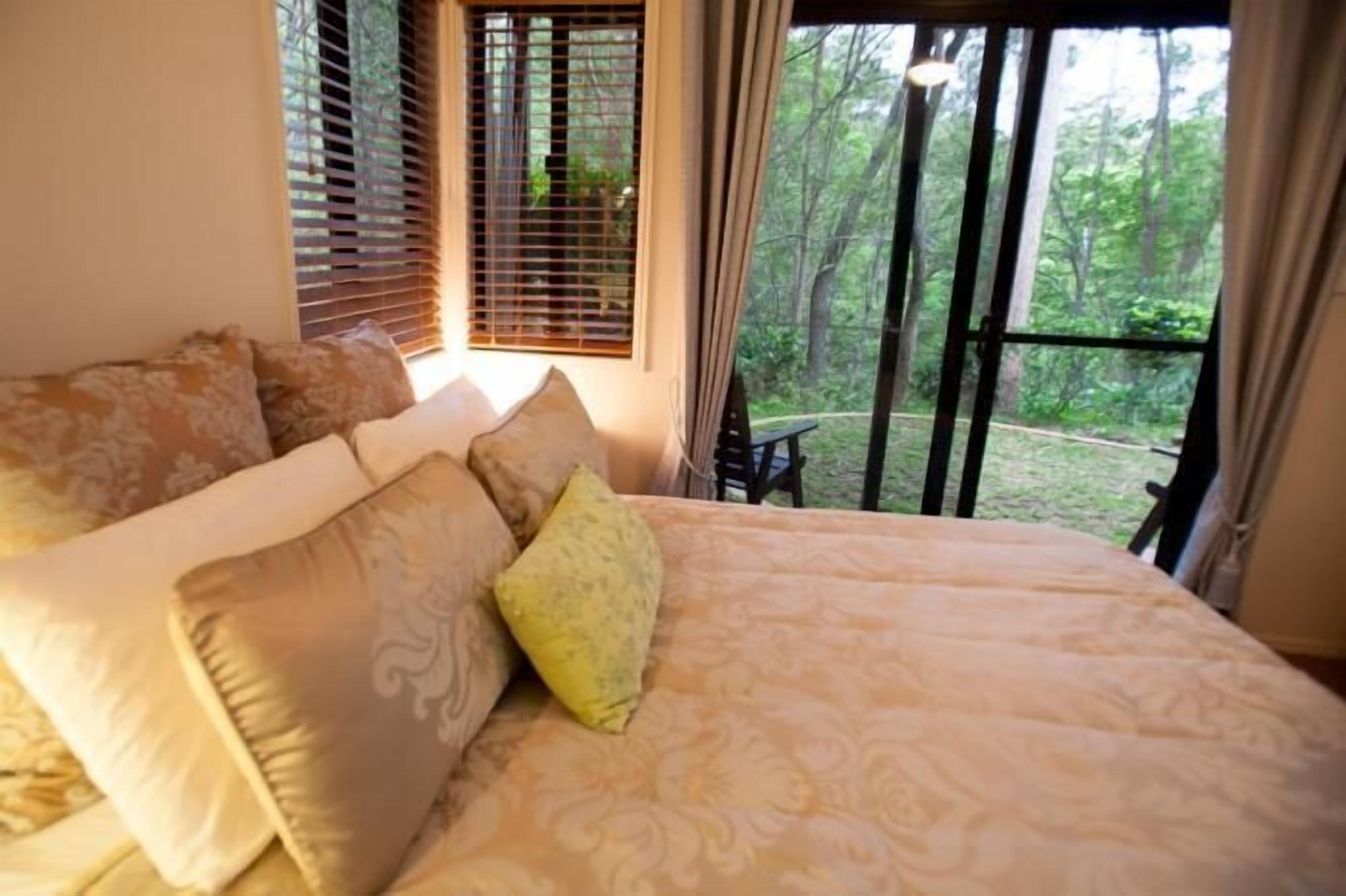 Quarto com cama grande, várias almofadas, janela com persianas e vista para árvores.