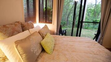 Quarto com cama grande, várias almofadas, janela com persianas e vista para árvores.