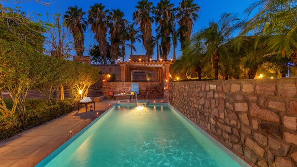 A swimming pool with stone walls, palm trees, and a seating area with a table and chairs. - Bijolai Palace - A Inde Hotel Jodhpur (Jodhpur)