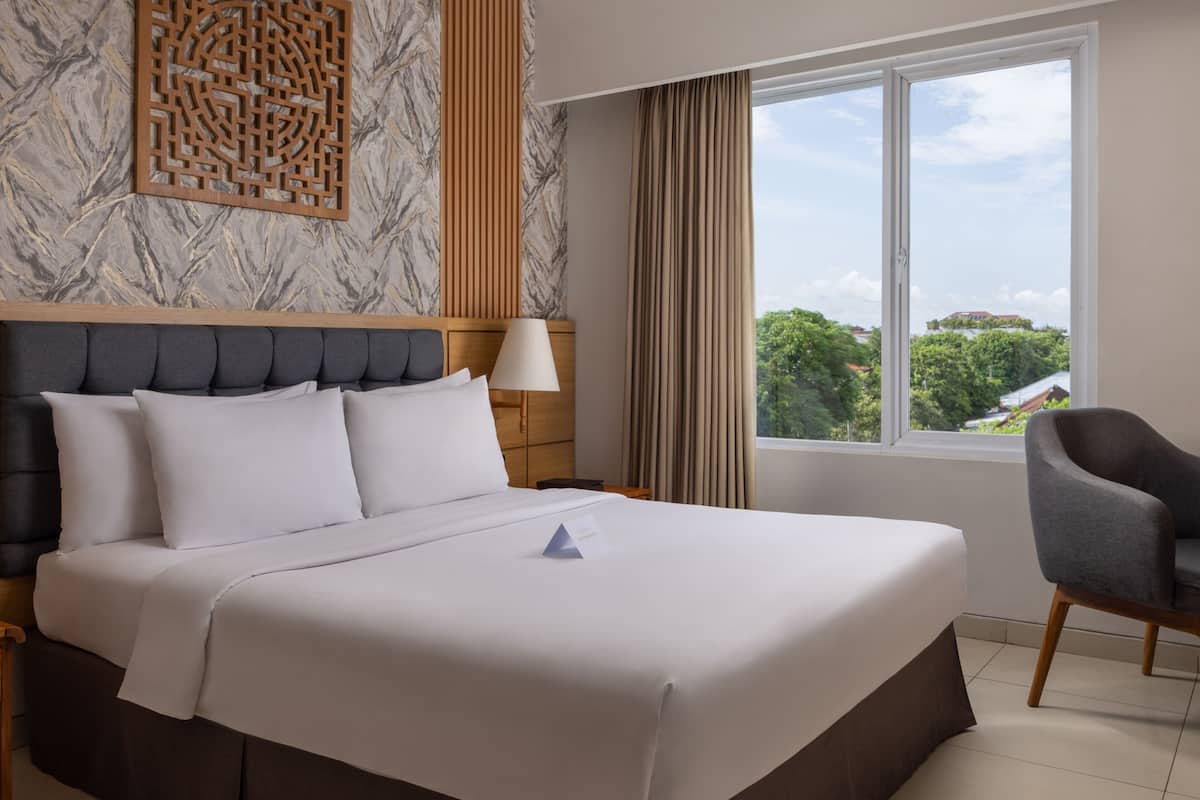 A hotel room with a large bed, a wooden headboard, a nightstand, a lamp, a chair, a window with a view, and a decorative wall panel.