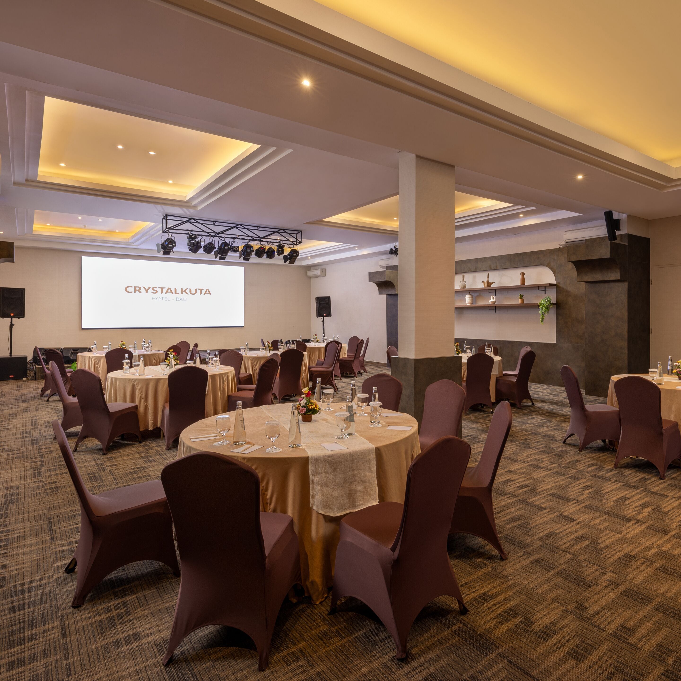 A conference room with round tables, chairs, a projector screen, and a stage with lighting equipment.