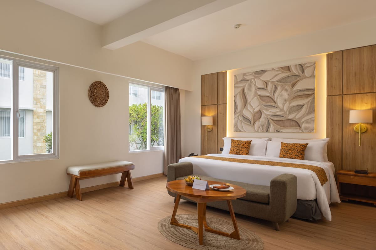 A hotel room with a large bed, a sofa, a round wooden table, and a view of greenery outside.