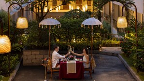 Un espace de restauration en plein air avec une table dressée pour deux personnes, entouré de verdure et illuminé de guirlandes lumineuses décoratives.