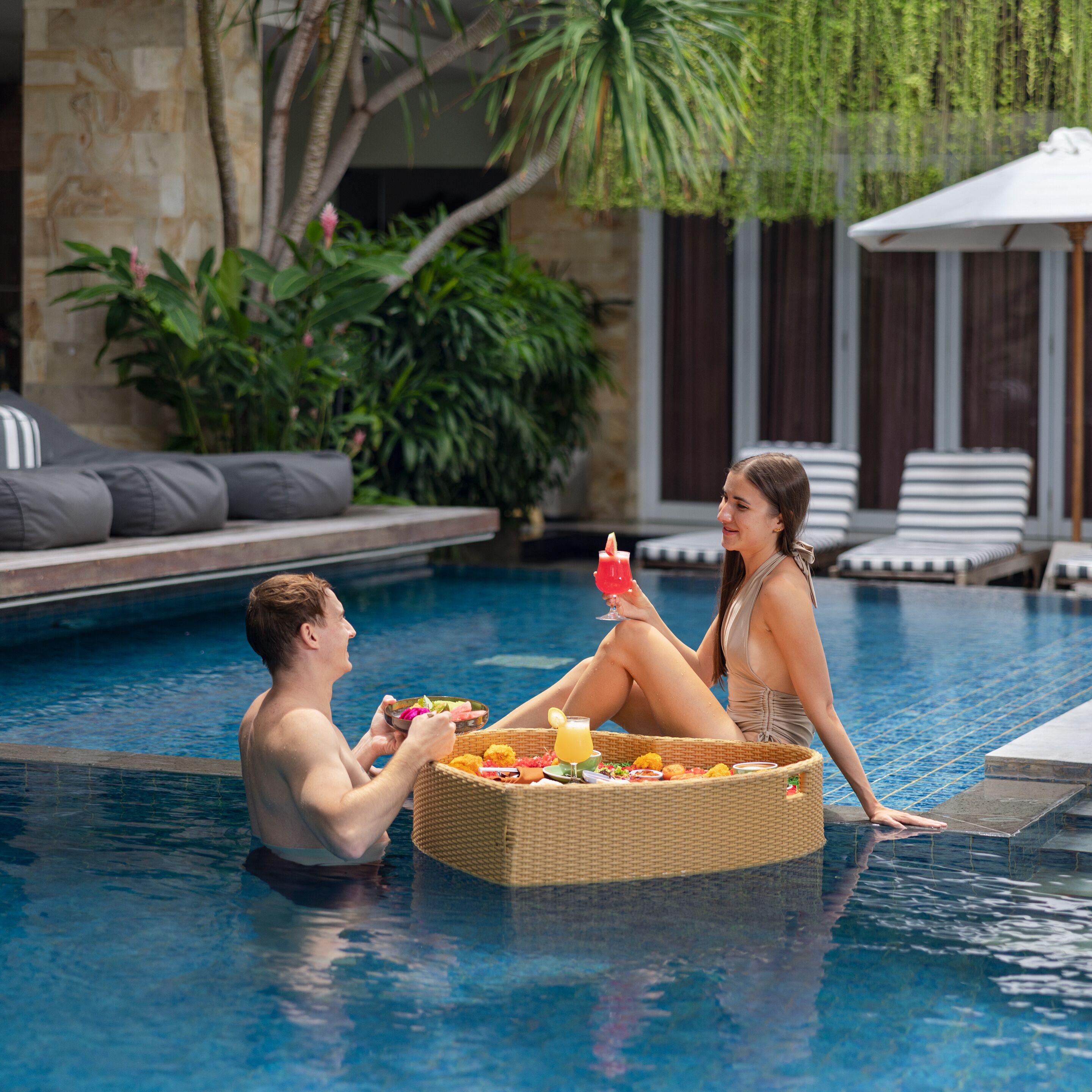 A couple relaxing by a poolside with a wicker basket of food and drinks.