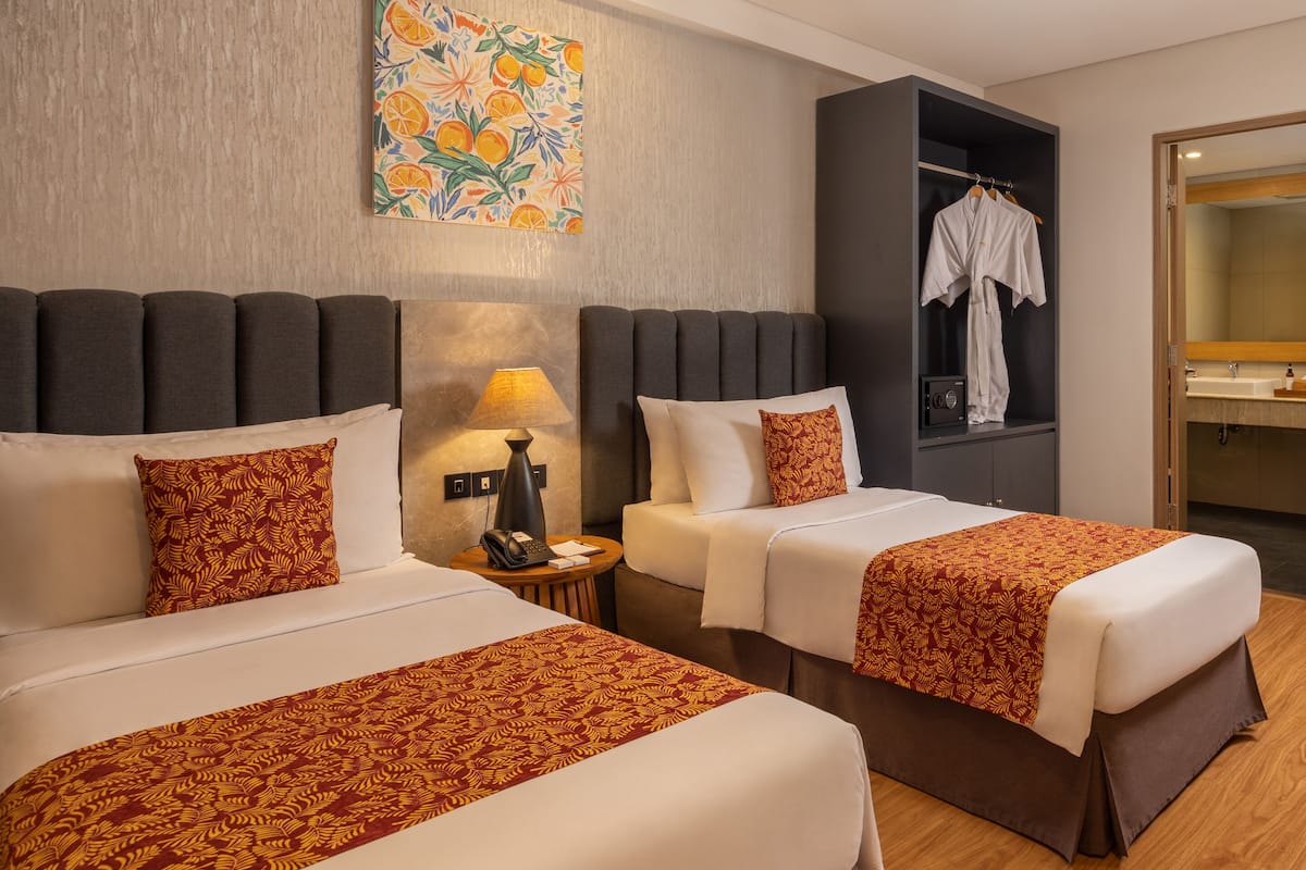 A hotel room with two beds, a nightstand, a lamp, a robe hanging, and a painting on the wall.