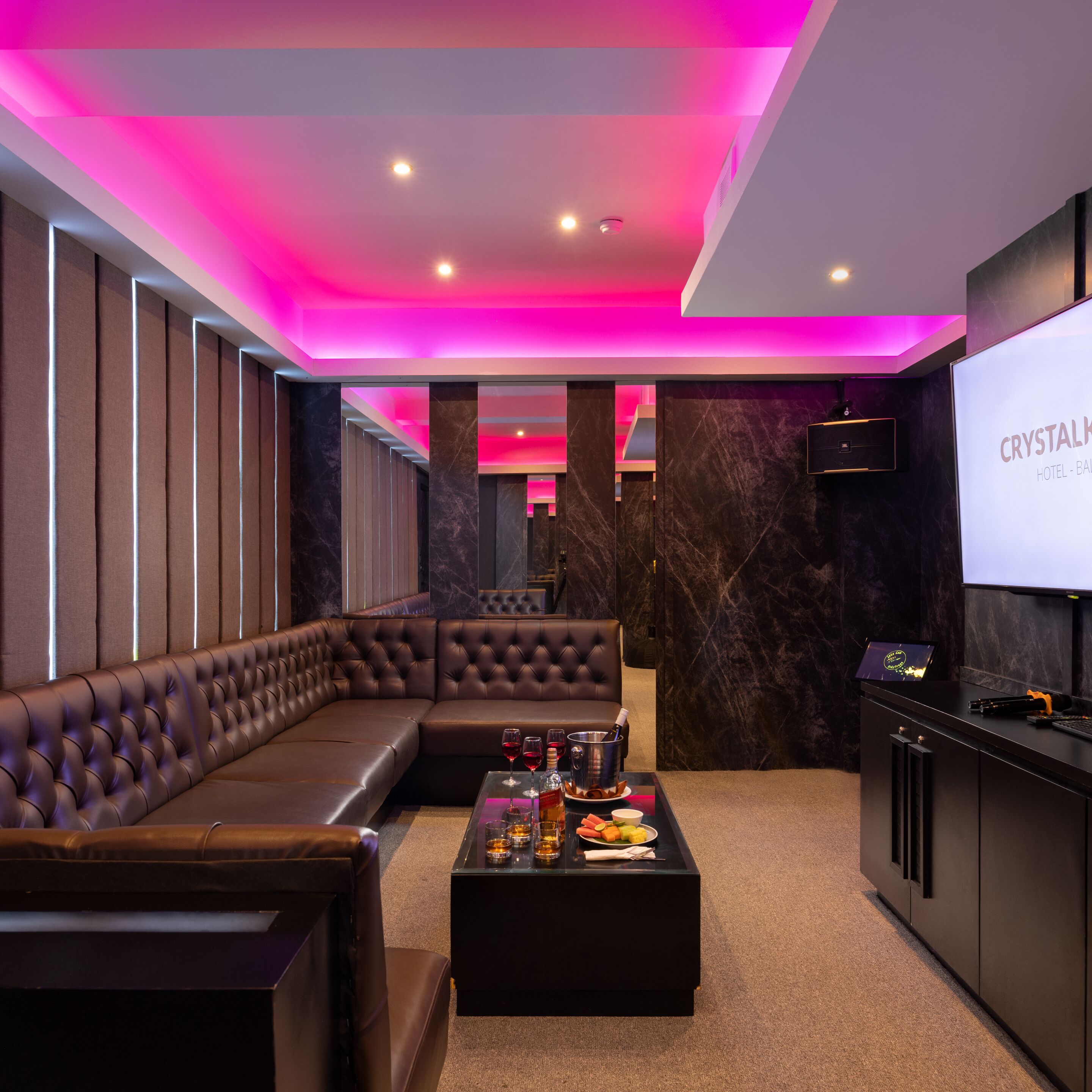 A modern hotel room with a leather sofa, a coffee table with snacks and drinks, and a TV screen displaying 'CRYSTALK HOTEL - BATHROOM'.