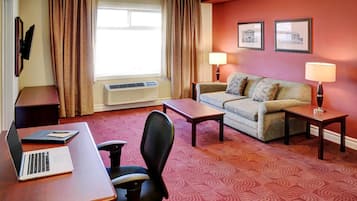 Ein Hotelzimmer mit Schreibtisch, Laptop, Stuhl, Sofa, zwei Nachttischen mit Lampen, einem Fenster mit Vorhängen und zwei gerahmten Bildern an der Wand.