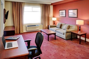 Ein Hotelzimmer mit Schreibtisch, Laptop, Stuhl, Sofa, zwei Nachttischen mit Lampen, einem Fenster mit Vorhängen und zwei gerahmten Bildern an der Wand.