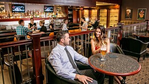 A bar area with multiple seating options, including bar stools and tables. There are several people seated and standing, and a bartender is visible in the background. - Chateau Nova Yellowhead (Edmonton)