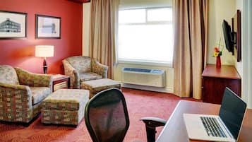 Habitación de hotel con dos sillones estampados, una mesita, una computadora portátil sobre un escritorio, una lámpara de pie, un televisor y una ventana con cortinas.