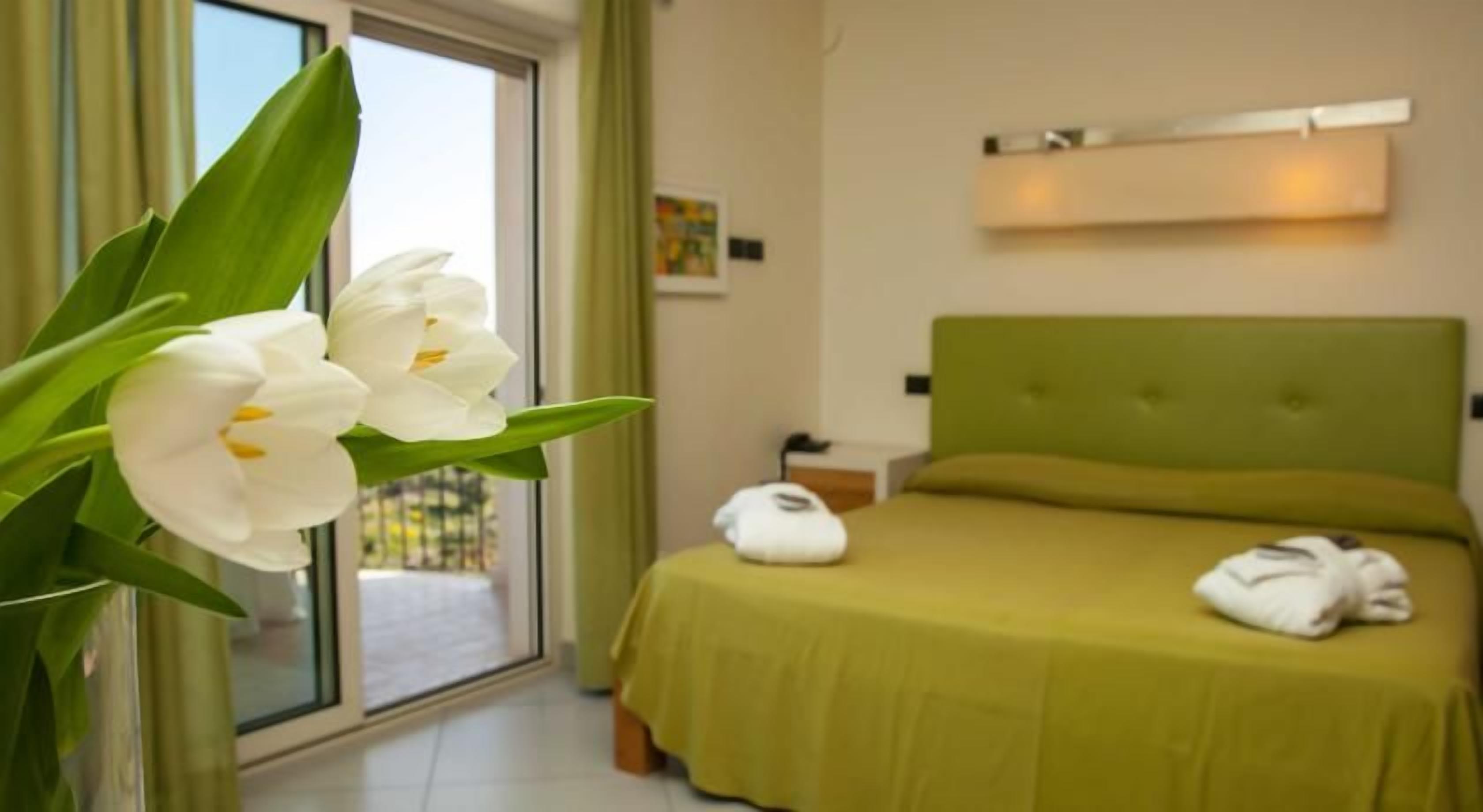 A hotel room with a bed, a green headboard, a window with a view, and a vase with white flowers.