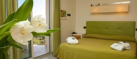 Ein Hotelzimmer mit Bett, grünem Kopfteil, Fenster mit Blick und einer Vase mit weißen Blumen.