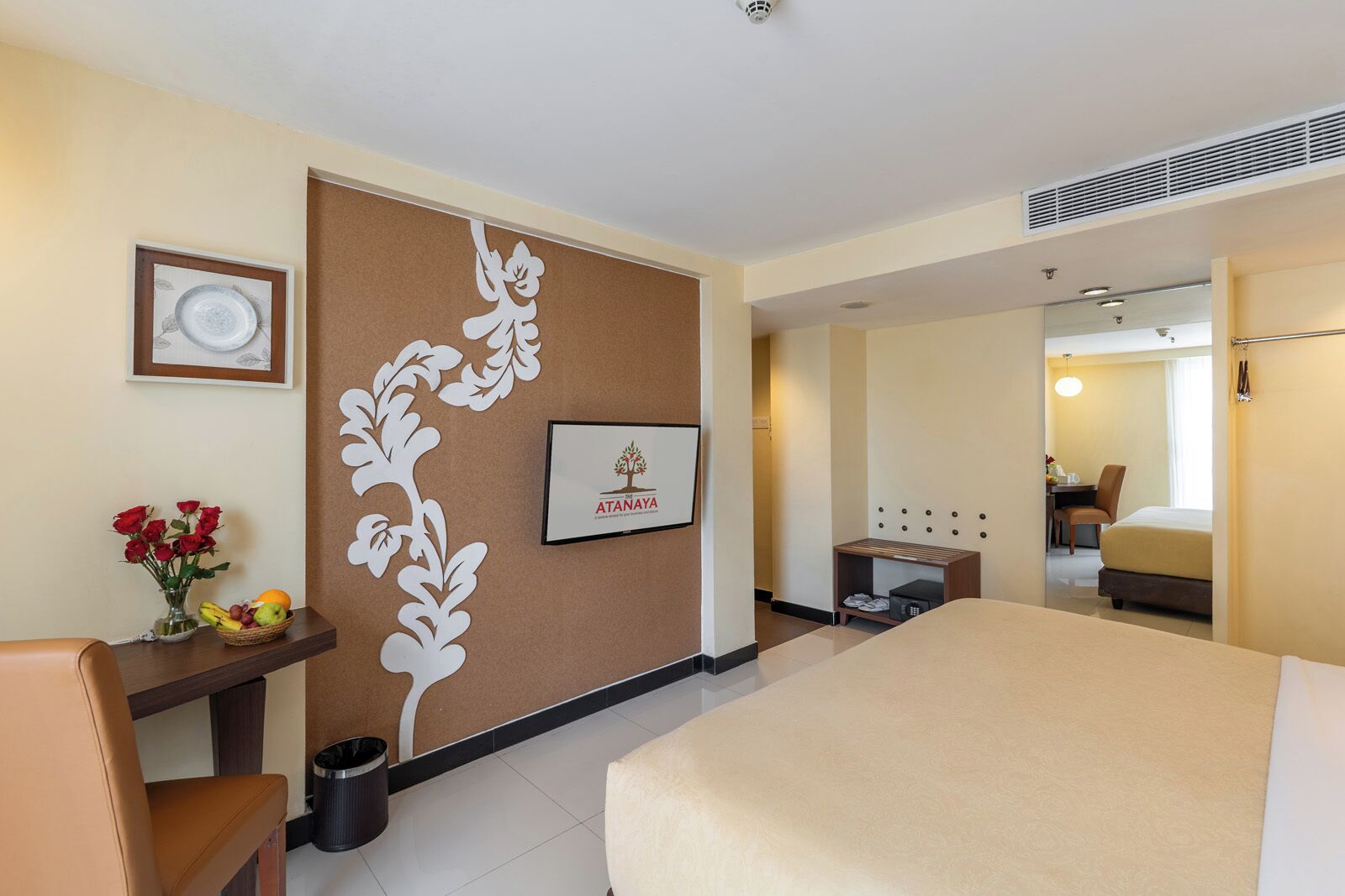 Habitación de hotel con cama, escritorio con silla, una pequeña mesa con silla, un cubo de basura, un cuadro enmarcado y un panel decorativo en la pared.