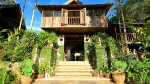 A rustic wooden house with a balcony, surrounded by lush greenery and potted plants.