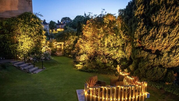 A well-lit garden area with a wooden deck, surrounded by lush greenery and trees.