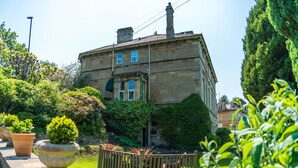 A historic building with a stone facade, large windows, and a garden with potted plants and a wooden fence.