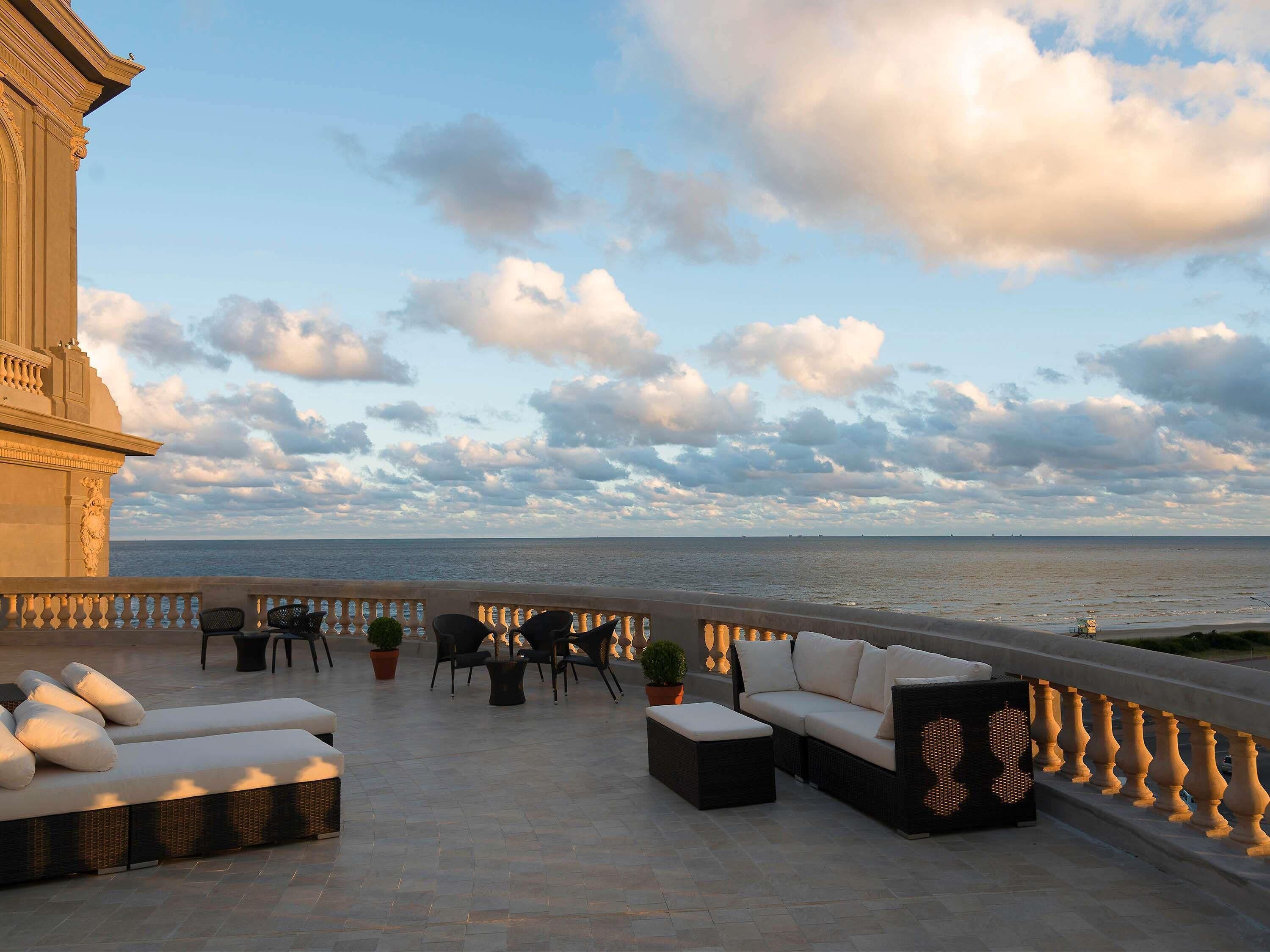 A terrace with outdoor seating, a view of the sea, and a building with classical architecture.