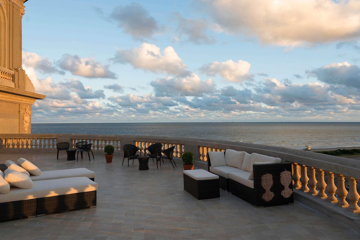 Una terraza con asientos al aire libre, vista al mar y un edificio de arquitectura clásica.