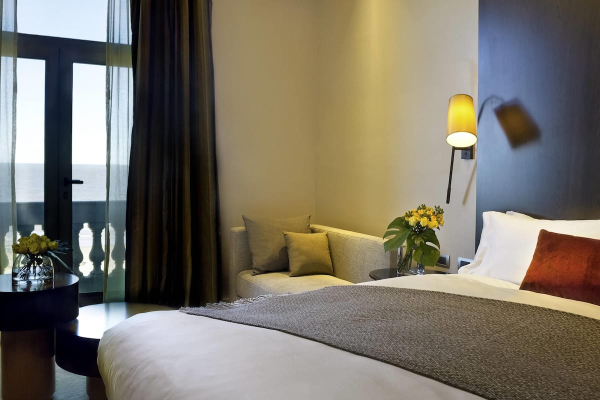 Ein Hotelzimmer mit Bett, Sofa, Nachttisch mit Lampe und Meerblick durch ein Fenster.