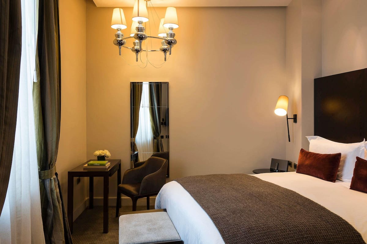 A hotel room with a large bed, a desk with a chair, a mirror, and a chandelier.