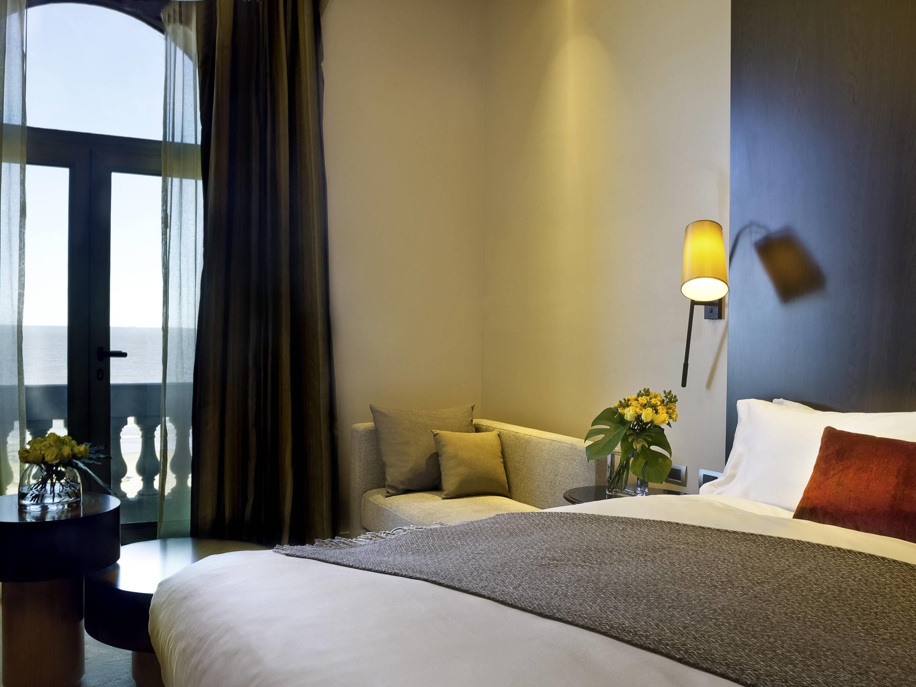 Ein Hotelzimmer mit Bett, Sofa, Nachttisch mit Lampe und Meerblick durch ein Fenster.