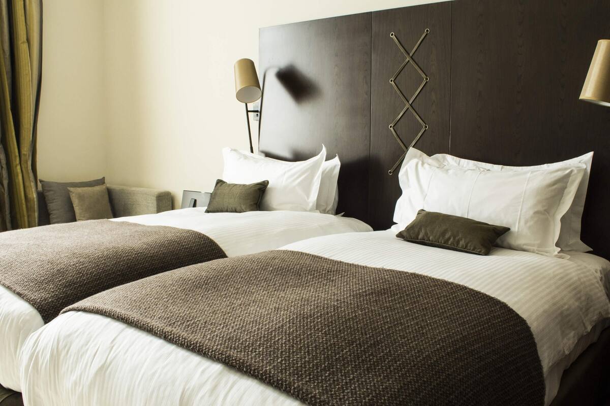 Une chambre d’hôtel avec deux lits, une tête de lit en bois foncé, du linge de lit blanc et deux lampes fixées au mur.